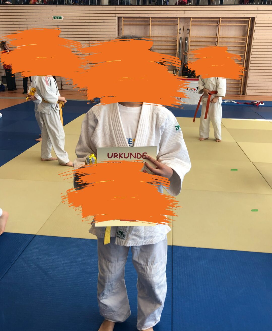 Ein Kind in einem weißen Judoanzug hält stolz eine Urkunde in den Händen. Im Hintergrund sind weitere Kinder in Judoanzügen auf einer Judomatte zu sehen. Die Gesichter der Personen wurden aus Datenschutzgründen unkenntlich gemacht.