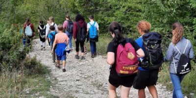 Eine Gruppe von Menschen wandert auf einem steinigen Weg durch eine grüne, bewaldete Landschaft. Einige tragen Rucksäcke, darunter ein auffälliger roter Rucksack mit der Aufschrift "Hogwarts". Der Weg führt leicht bergauf, und im Hintergrund sind Hügel und blauer Himmel zu sehen.
