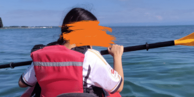 Eine Person sitzt in einem roten Kajak und paddelt auf einem ruhigen Gewässer unter blauem Himmel. Sie trägt eine rote Schwimmweste und hält ein Paddel in der Hand. Im Hintergrund sind das Ufer und einige Bäume sowie ein weitläufiger Horizont zu sehen.