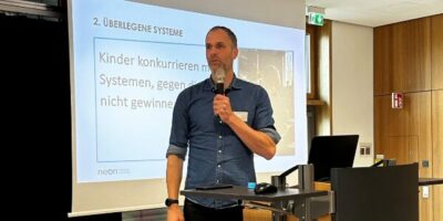 Ein Mann hält einen Vortrag und steht hinter einem Rednerpult in einem modernen Konferenzraum. Im Hintergrund ist eine Präsentationsfolie mit der Überschrift „Überlegene Systeme“ zu sehen. Auf dem Boden vor dem Pult steht eine Wasserflasche, und die Beleuchtung im Raum ist hell.