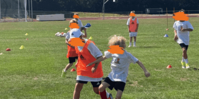 Mehrere Kinder spielen bei sonnigem Wetter Fußball auf einem grünen Rasenplatz während eines Trainings. Im Vordergrund kämpfen zwei Jungen intensiv um den Ball, wobei einer ein orangefarbenes Leibchen und der andere ein weißes T-Shirt trägt. Im Hintergrund sind weitere Mitspieler in Sportkleidung, Trainingshütchen, Tore und eine dichte Baumreihe zu sehen.