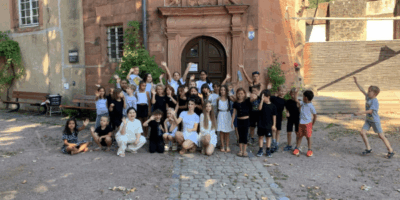 Eine große Gruppe von Kindern und Betreuern posiert fröhlich auf dem gepflasterten Innenhof vor dem Eingang eines historischen Gebäudes. Einige Kinder knien vorne, während die anderen dahinter stehen und jubelnd die Hände in die Luft strecken. Das Bild zeigt die Gemeinschaft und gute Stimmung aller Teilnehmenden des Musikcamps im Freien.