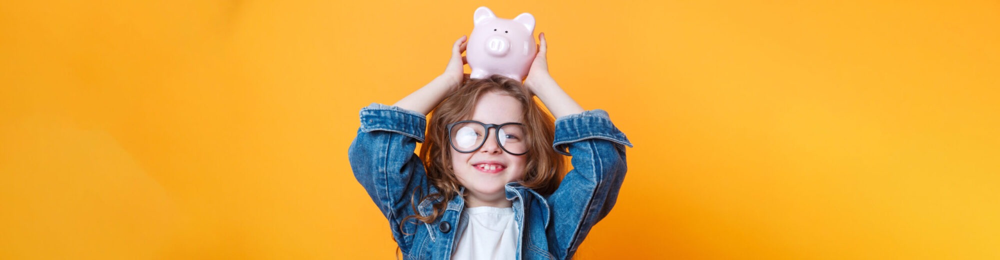 Ein Kind in einer Jeansjacke hebt ein rosa Sparschwein über seinen Kopf. Der Hintergrund ist leuchtend orange und erzeugt eine fröhliche Atmosphäre. Das Bild vermittelt eine positive Botschaft über Sparen und finanzielle Bildung für Kinder.
